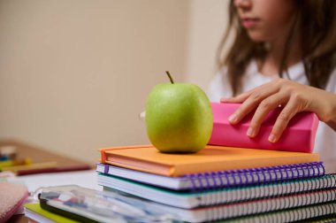 Bir sınıf sahnesinde bir öğrencinin masası yeşil bir elma ile renkli defterlerin üzerinde duruyor, okul malzemelerinin yanında kalem kutusu gibi. Akademik ortamlarda eğitim, öğrenme ve organizasyonu vurguluyor.