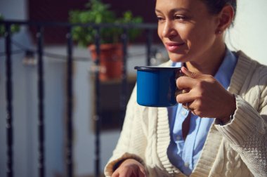 Mavi kupa tutan bir kadının açık havada içkinin tadını çıkardığı huzurlu bir an. Sahne, onun ifadesini aydınlatan yumuşak güneş ışığı tarafından vurgulanan rahatlama, temiz hava ve sıcaklığı yakalıyor..