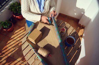 Dekoratif kahverengi kağıt, makas ve sicimle tahta bir masaya hediye paketleyen bir kadın. Ortam güneşli, saksı bitkileri görünür, rahat ve yaratıcı bir atmosfer yaratıyor..