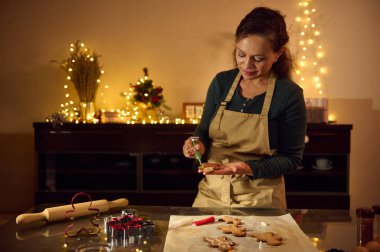 Önlük takan bir kadın, Noel ışıklarıyla süslenmiş sıcak bir mutfakta zencefilli kurabiye süslüyor. Sette bayram neşesi yayılıyor, tatil pişirme ve yaratıcı şenliklerin sevinci vurgulanıyor.