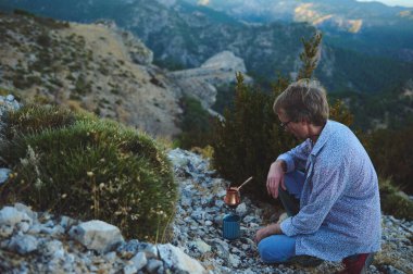 Bir adam sakin bir dağ ortamında portatif bir sobayla kahve hazırlıyor. Çevrenin doğal güzelliği sakin atmosferi güçlendirir. Açık hava rahatlama ve yaşam tarzı tasviri için uygun.
