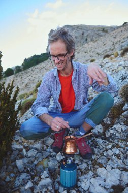 Bir adam güneşli bir gökyüzünün altındaki kayalık bir dağ bölgesinde taşınabilir kamp sobası kullanarak yemek hazırlıyor. Rahat duruşu ve engebeli yapısı maceraperest ve sakin doğa yaşamını vurguluyor..