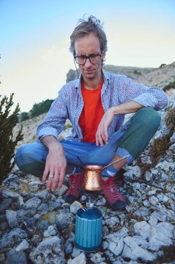 Bir adam, kayalık arazide doğal bir şekilde oturup taşınabilir gaz sobası kullanarak sıcak bir içecek hazırlıyor. Günlük kıyafetleri sakin açık hava ortamını tamamlıyor, sükunet ve macera sergiliyor..