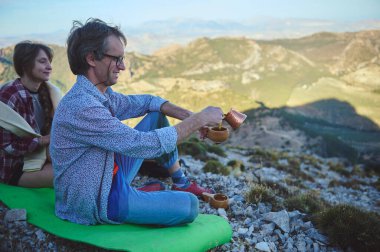 İki kişi dağlık bir arazide çimenli bir yamaçta oturur, biri geleneksel bir çaydanlıktan bir fincana kahve doldurur. Görüntü, sakin bir rahatlama anı ve açık havanın takdir edilmesini resmediyor..