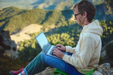 Dağlık engebeli bir arazide oturan bir insan dizüstü bilgisayarla çalışır. Sakin doğal ortam ve manzaralı zemin, uzak çalışma, özgürlük ve sükunet kavramlarını simgeliyor..