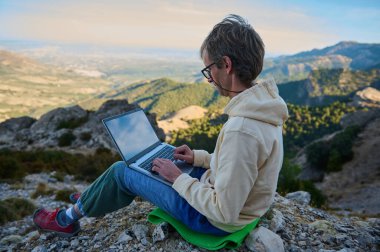 Doğal bir dağ ortamında dizüstü bilgisayarda bireysel tipler, özgürlüğü, üretkenliği ve modern teknolojiyi vurguluyor. Bu sahne, açık havada esnek çalışma ortamlarının özünü yakalar..