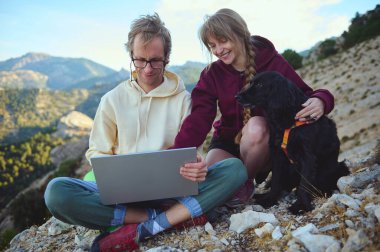 Bir çift kayalık bir yamaçta birlikte oturur ve evcil köpekleri dikkatle izlerken dizüstü bilgisayarda çalışırlar. Dekor, güzel doğal çevresi olan manzaralı, dağlık bir alanı vurguluyor..