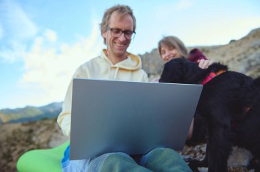 Gülümseyen çift, doğayla iç içe olan bir bilgisayarda çalışıyor ve yanında köpekleri de var. İlham verici bir ortamda boş vakitleri ve verimliliği somutlaştırıyorlar..