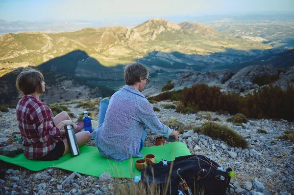 İki arkadaşın dağın yamacında oturup aşağıdaki panoramik manzarayı izlerken yeşil bir hasırda dinlendikleri huzurlu bir an. Macera, rahatlama ve doğa ile bağlantı kavramları için mükemmel..