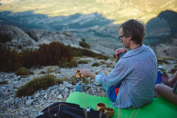 Bir adam yamaçta otururken taşınabilir bir düzenek kullanarak kahve hazırlıyor. Etrafı kayalık araziyle çevrili ve uzak manzaralı manzarayla çevrili sakin, açık hava macerasının tadını çıkarıyor..