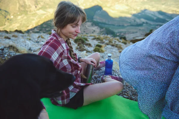 Ekose gömlekli genç bir kadın dışarıda bir tepede oturuyor. Yanında başkaları ve siyah bir köpek var. Rahatlatıcı ve bağlayıcı bir atmosfer yaratıyor. Yakında bir şişe su ve matara var..