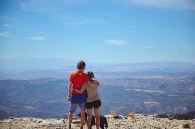 Bir çift, bir dağın zirvesinde durmuş vadinin ve uzak dağların geniş manzarasının tadını çıkarıyorlar. Açık mavi gökyüzü nefes kesen panoramik manzarayı vurgular..
