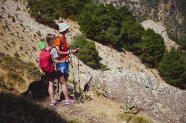 İki yürüyüşçü ve köpekleri, doğa ve maceranın tadını çıkararak, pitoresk bir dağ yolunda ilerliyorlar. Sırt çantaları ve yürüyüş direkleri vahşi doğada tatmin edici bir deneyime hazırlanıyor..