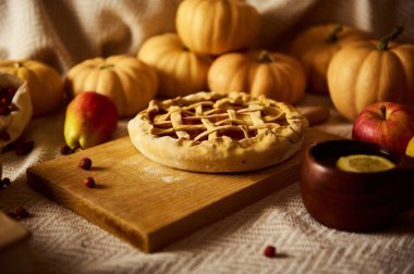 Ahşap bir tahtanın üzerinde kafes şeklinde bir balkabağı turtası, ahşap bir kasede limon dilimi ve balkabağı ve elmalarla çevrili bir sonbahar sahnesi Şükran Günü sıcaklığını çağrıştırıyor..