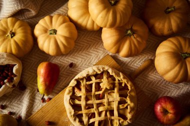 Balkabakları, elmalar ve ahşap bir tahtanın üzerinde örülü bir turtanın yer aldığı rahat bir sonbahar hayatı. Ilık ışıklandırma ve örülmüş kumaş perde arkası Şükran Günü sıcaklığı ve mevsimsel hasat titreşimleri yaratır..
