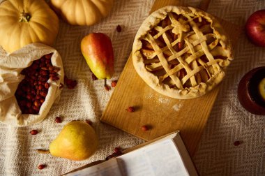 Ahşap bir tahtanın üzerinde kafes turtası, balkabağı, elma, armut ve kurutulmuş yaban mersini içeren rahat bir sonbahar hayatı. Açık bir kitap, sıcak Şükran Günü havasını tamamlar. Ev yapımı anları, hasat tatlarını ve mevsimsel kutlamaları davet eder..