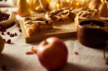 Autumn table scene with sliced pie on a wooden board, apples, a dark mug, and warm fairy lights. Cozy holiday mood perfect for Thanksgiving, home cooking, and seasonal celebrations.