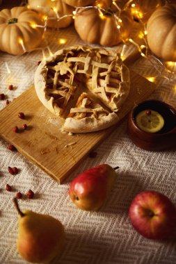 Ahşap bir tahtanın üzerinde kafes kabuklu bir turta, balkabağı, elma, armut ve dağınık baharatların olduğu rustik bir sonbahar masası sahnesi. Sıcak peri ışıkları parıldıyor. Meyve, sade doku ve mevsimsel renklerle, Şükran Günü havası yaratıyor..