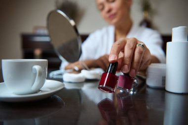 Kaplıca ortamındaki bir kadın iki farklı oje renginde seçer. Şişeler ve kavanozlar gibi bir fincan taze kahve ve kişisel bakım ürünleri huzurlu ve rahatlatıcı atmosferi tamamlıyor.