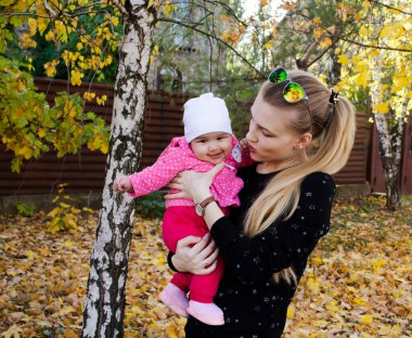 Genç ve güzel bir kadın gülen bebekle sonbahar parkında konuşuyor. Sarı ağaçların altında kızını kucaklayan genç bir anne. Sarışın kadın 3 aylık bebeği kollarında tutuyor. Aile doğayı sever.