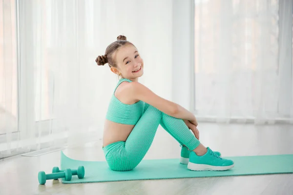 Маленька спортивна дівчинка гімнастка в спортивному одязі виконує вправи на килимку в приміщенні Стокове Зображення