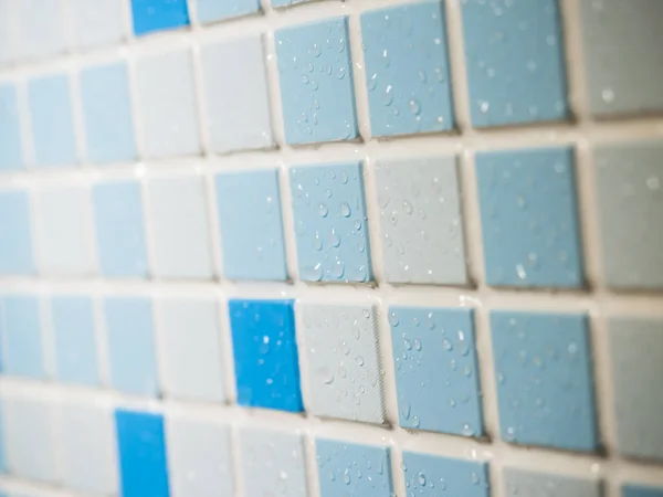 A wall of blue, blue and white small tiles with water droplets on the surface, at an angle.