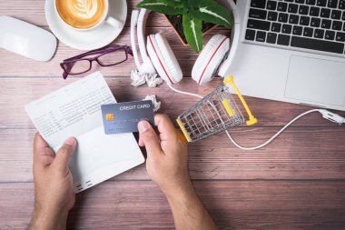 Adamın biri bir kredi kartı tutuyor ve tahtadan ofis masasındaki hesap defterinden bakiyesini kontrol ediyor. Laptop, kulaklık ve sıcak kahveden oluşuyor. İnternetten alışveriş kavramı.