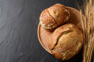 Kırsal ahşap bir tabakta sunulan taze pişmiş, kabuklu, ekşi ekşi ekmeğin üst görüntüsü. Tırtıklı bir bıçak ekmeğin yanında duruyor, bir demet altın buğday kulağı kenarda zarif bir şekilde duruyor, hepsi koyu taştan bir arka planda duruyor..