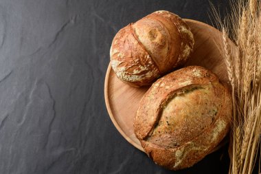 Kırsal ahşap bir tabakta sunulan taze pişmiş, kabuklu, ekşi ekşi ekmeğin üst görüntüsü. Tırtıklı bir bıçak ekmeğin yanında duruyor, bir demet altın buğday kulağı kenarda zarif bir şekilde duruyor, hepsi koyu taştan bir arka planda duruyor..
