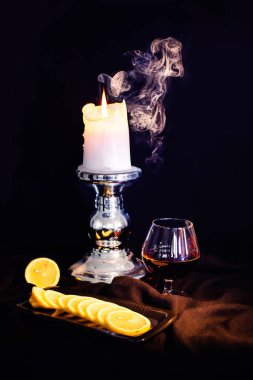 Bir bardak konyak, yanan bir mum ve dumanın arka planında bir tabakta limon.