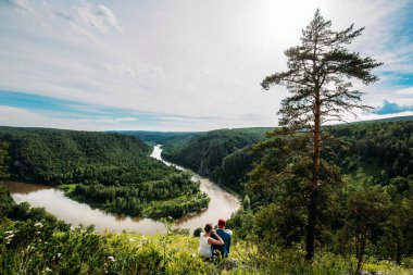 Çift oturur ve Nehri yakınında güzel bir gün batımı bakar. Erkek ve kadın nehirde bak ve doğanın tadını çıkarın. Güzel çift erkek ve sarılma ve rahatlatıcı nehir oturan kadın arkadan görünüm.