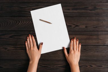 Kadınların ellerinin üst görünümü, tahta bir masa üzerinde yatan boş bir kağıt parçası üzerinde bir şeyler yazmaya hazır. Ahşap arka plan üzerinde kalem ile kağıt beyaz boş levha. Beyaz bir sayfaya mektup yazma