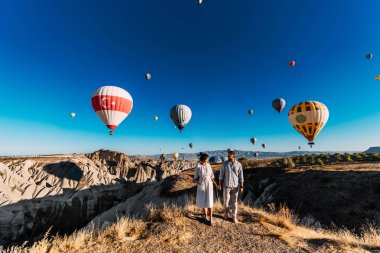 Aşık bir çift, Cappadocia 'da balonların arka planında duruyor. Çift dünyayı geziyor. Türkiye 'de tatil. Balayı gezisi. Sıcak hava balonu uçuşları. Mutlu genç çift Kapadokya 'da şafakla tanışır.