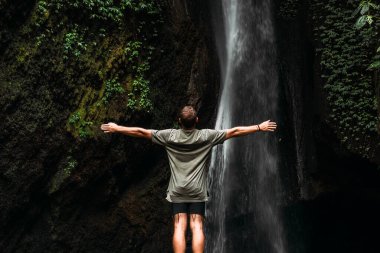 Şelalenin yanında kolları açık duran bir adamın arka görüntüsü. Şelaledeki adam doğaya daha yakın hissetmek için ellerini kaldırıyor. Şelaledeki adam. Bali 'ye git. Doğanın arka planındaki adam.