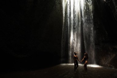 Beautiful couple in a cave with a waterfall. Athletic man and woman under the streams of a waterfall. A sexy couple under a tropical waterfall in twilight light. Tukad Chepung Waterfall. Copy space clipart