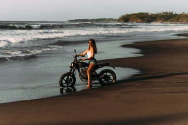 Sahilde motosikletli güzel bekar kız gün batımıyla buluşuyor. Deniz kenarında bisikleti olan şık bir kadın. Motosikletle seyahat et. Bali 'ye git. Tropikal plajda özel bir motosiklet. Boşluğu kopyala