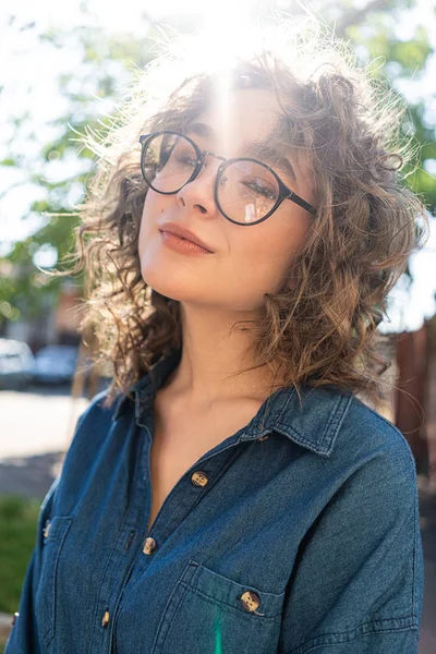 Outdoor close up portrait of young beautiful stylish happy smiling curly girl wearing sunglasses, posing in street. Sunny day light. Summer fashion concept. Copy, empty space for text