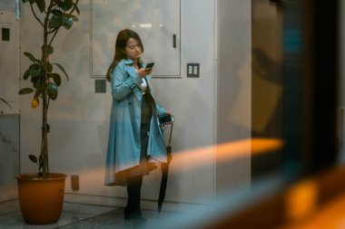 Pencerenin arkasında cep telefonu olan mavi ceketli bir kız..