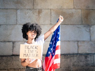 Amerikan bayrağı ve siyah bir hayat işareti taşıyan genç siyahi kadın izole edilmiş, açık havada, arka planda taş duvar.