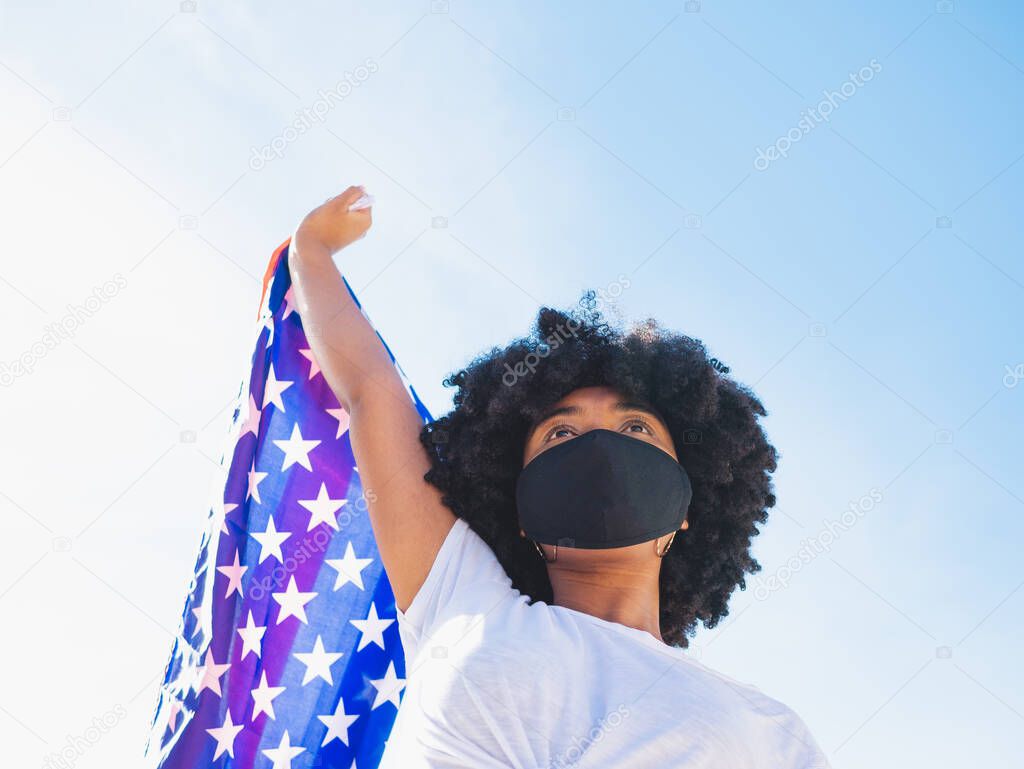 Portrait of a young black woman wearing a protective mask and holding a USA flag, outdoors, isolated, with copy space