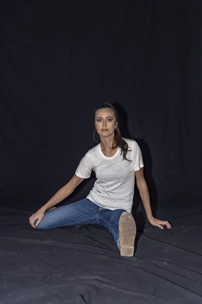 Ragazza in jeans blu e t-shirt bianca seduta sul pavimento. Fondo nero — Foto Stock