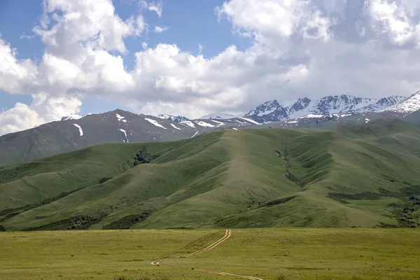 Estrada rural entre campos verdes e colinas contra o pano de fundo das montanhas com picos cobertos de neve e um céu nublado . — Fotografia de Stock