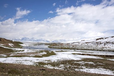 Otlaklar arasındaki nehir karla kaplı. Ufukta bir dağ sırası var. Bulutlarda mavi gökyüzü