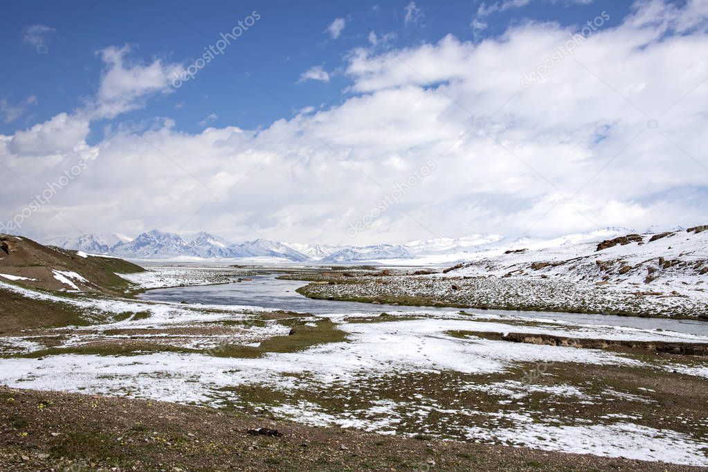 The river between pastures covered with snow. On the horizon is a mountain range. Blue sky in the clouds