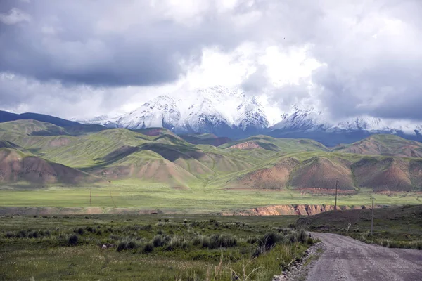 Onverharde weg tussen bloeiende velden en pittoreske bergketens aan de horizon. Kirgizië Naryn Region — Stockfoto