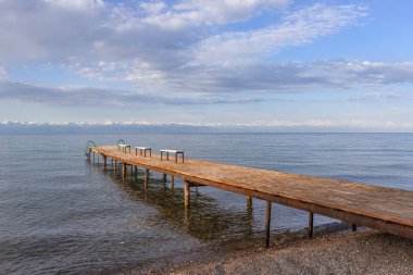 Issyk-Kul Gölü 'nde sıralı rıhtım, ufukta bulutlu bir gökyüzüne karşı karla kaplı tepeler var.