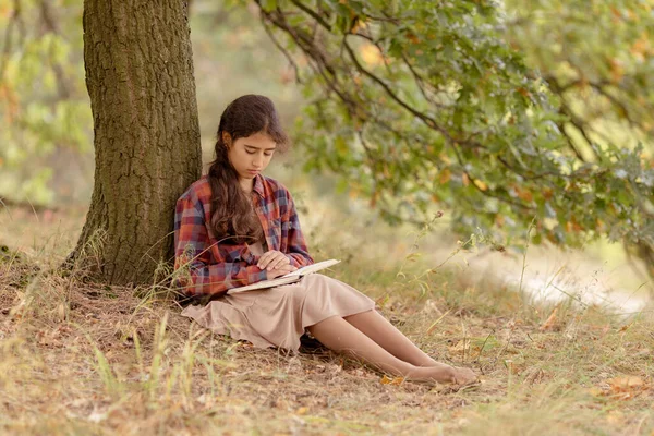 Girl intently reads a book under a tree in the park.