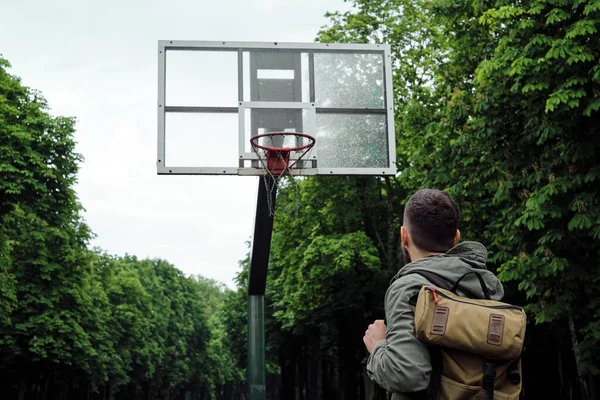 A man standing in front of a basketball basket. Focused on the goal, looking at the basketball basket. Street basketball. Walk in the park with a backpack on a rainy day. Urban lifestyle, outdoor