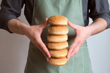 Bir adamın elinde hamburger çörekleri. Çörek pişirmek. Hamburger, yemek için malzeme. Önlüklü bir şef. Susam tohumlu güzel taze çörekler. Geleneksel Amerikan Lezzetli Yemeği