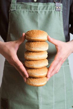 Bir adamın elinde hamburger çörekleri. Çörek pişirmek. Hamburger, yemek için malzeme. Önlüklü bir şef. Susam tohumlu güzel taze çörekler. Geleneksel Amerikan Lezzetli Yemeği